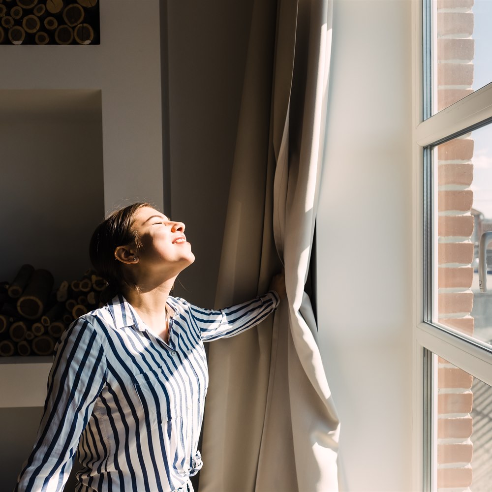 Woman smiling by window opening curtains in sunlight